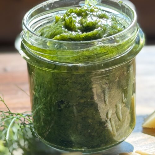 vasetto di pesto appoggiato a tagliere in legno con barbe di finocchio pezzi di formaggio e pinoli