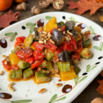Caponata di ottobre con melanzane, zucchine, peperoni e pomodorini al forno, completata con capperi, pinoli tostati ed erbette aromatiche.