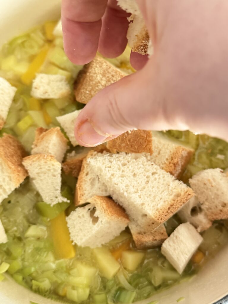 Mani che spezzano e aggiungono il pane raffermo nel tegame di ghisa per preparare la Panada con pane raffermo.
