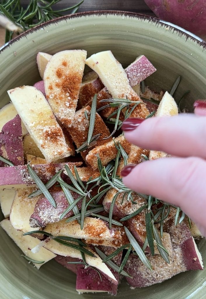 Mani che aggiungono rosmarino alle batate rosse tagliate a spicchi.